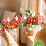 A person holds a paper Halloween banner spelling 'trick or treat' in red-orange letters, decorated with cartoon ghosts, pumpkins, and flowers. The scene is set in a warm, softly blurred bedroom environment with a green 'Glow in the Dark' badge overlay in the bottom right corner.
