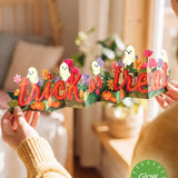 A person holds a paper Halloween banner spelling 'trick or treat' in red-orange letters, decorated with cartoon ghosts, pumpkins, and flowers. The scene is set in a warm, softly blurred bedroom environment with a green 'Glow in the Dark' badge overlay in the bottom right corner.