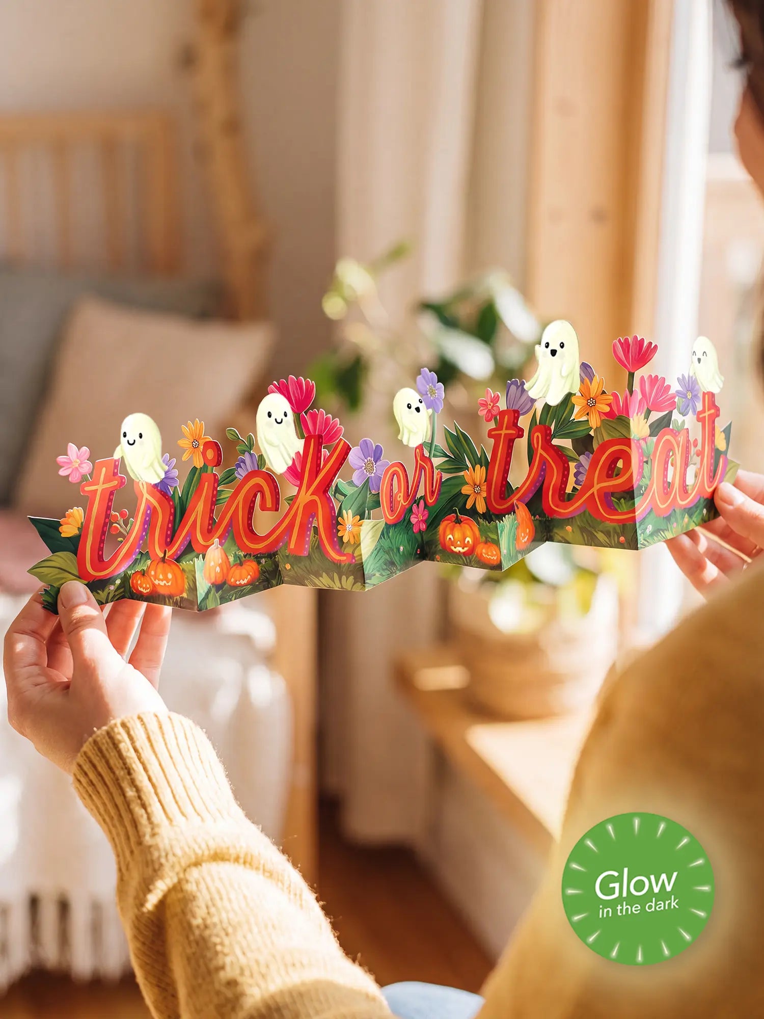 A person holds a paper Halloween banner spelling 'trick or treat' in red-orange letters, decorated with cartoon ghosts, pumpkins, and flowers. The scene is set in a warm, softly blurred bedroom environment with a green 'Glow in the Dark' badge overlay in the bottom right corner.