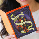 A smiling woman holds an open Halloween pop-up card. The card features a dark purple ornate frame with a 3D scene of a haunted house, ghosts, and pumpkins, wrapped with a light green banner that reads 'Happy Halloween!'.