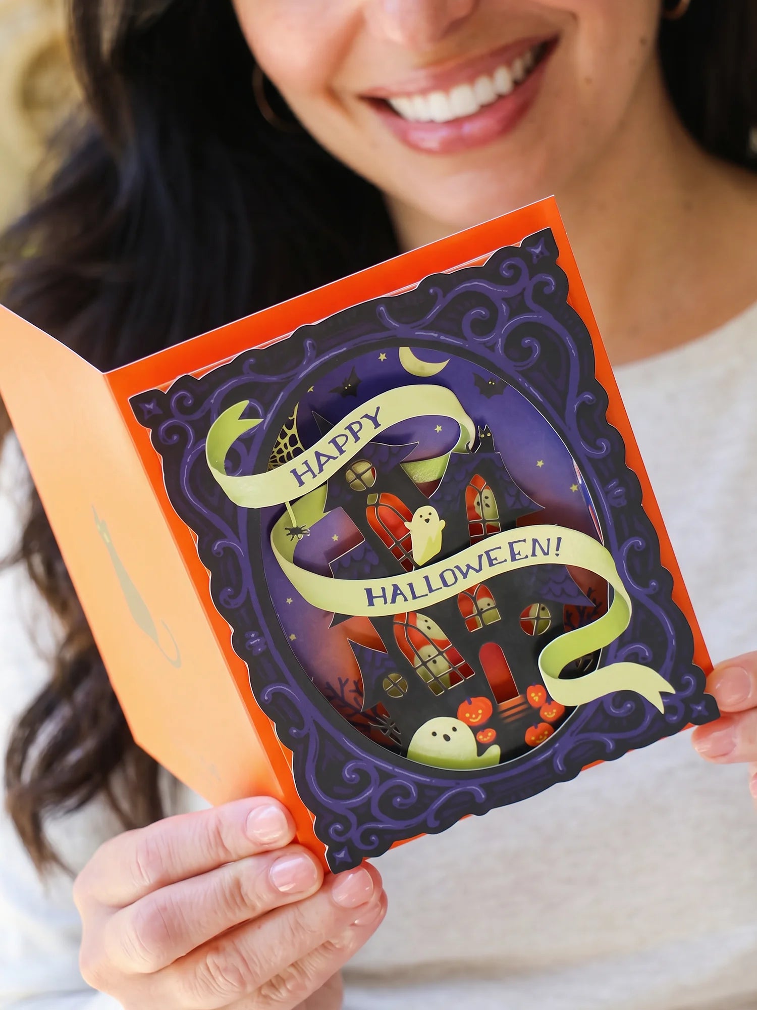 A smiling woman holds an open Halloween pop-up card. The card features a dark purple ornate frame with a 3D scene of a haunted house, ghosts, and pumpkins, wrapped with a light green banner that reads 'Happy Halloween!'.