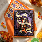 A Halloween greeting card with a spooky haunted-house illustration layered cut-out and a cream ribbon reading “Happy Halloween!” layered over an orange envelope with a pumpkin-and-bat pattern. The card is styled on a textured white fabric with orange flowers, candy corn, and a green “Glow in the Dark” badge.
