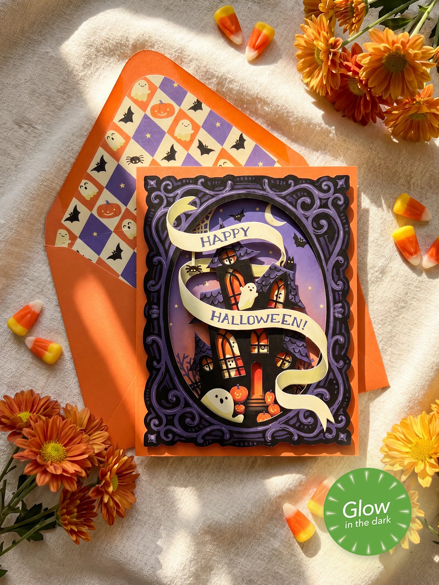 A Halloween greeting card with a spooky haunted-house illustration layered cut-out and a cream ribbon reading “Happy Halloween!” layered over an orange envelope with a pumpkin-and-bat pattern. The card is styled on a textured white fabric with orange flowers, candy corn, and a green “Glow in the Dark” badge.