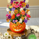 A Halloween-themed floral centerpiece in a carved orange pumpkin pot, filled with colorful artificial flowers and ghost decorations, displayed on a wooden stand on a table. A green circular badge reads 'Glow in the dark,' and small pumpkins and white daisies decorate the scene.