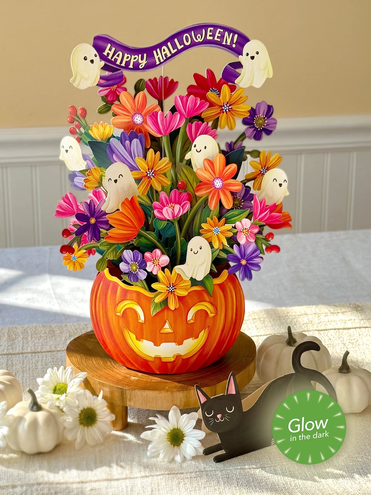 A Halloween-themed floral centerpiece in a carved orange pumpkin pot, filled with colorful artificial flowers and ghost decorations, displayed on a wooden stand on a table. A green circular badge reads 'Glow in the dark,' and small pumpkins and white daisies decorate the scene.