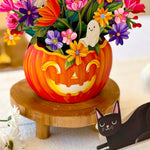 A Halloween-themed floral arrangement in a carved orange pumpkin vase, displayed on a wooden stand. The arrangement features colorful paper flowers, ghost cutouts, and green leaves, with a paper black cat figurine nearby on a light fabric surface.