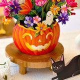A Halloween-themed floral arrangement in a carved orange pumpkin vase, displayed on a wooden stand. The arrangement features colorful paper flowers, ghost cutouts, and green leaves, with a paper black cat figurine nearby on a light fabric surface.
