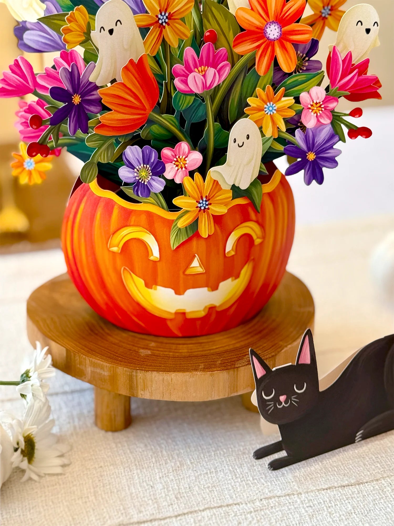 A Halloween-themed floral arrangement in a carved orange pumpkin vase, displayed on a wooden stand. The arrangement features colorful paper flowers, ghost cutouts, and green leaves, with a paper black cat figurine nearby on a light fabric surface.