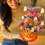 A woman in a beige sweater and blue jeans holds a colorful Halloween flower arrangement in a carved orange pumpkin pot. The arrangement features paper flowers, small white ghost cutouts, and a purple banner that reads 'Happy Halloween!'.