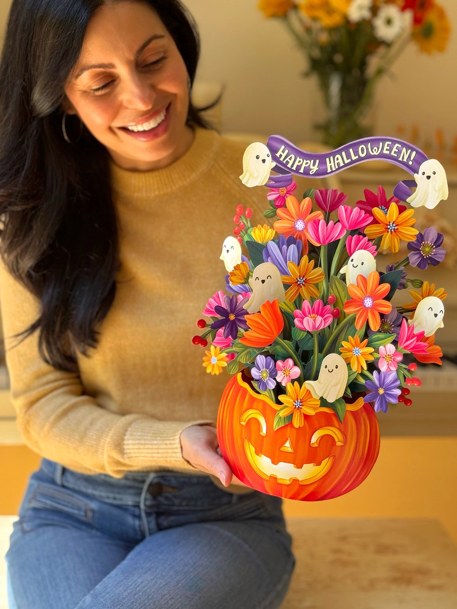 A woman in a beige sweater and blue jeans holds a colorful Halloween flower arrangement in a carved orange pumpkin pot. The arrangement features paper flowers, small white ghost cutouts, and a purple banner that reads 'Happy Halloween!'.