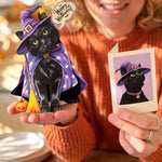 A woman in an orange sweater holds a black cat pop-up card dressed as a witch in a purple hat and cape with a broom, alongside a small 'Happy Halloween' card and a printed photo of the cat.