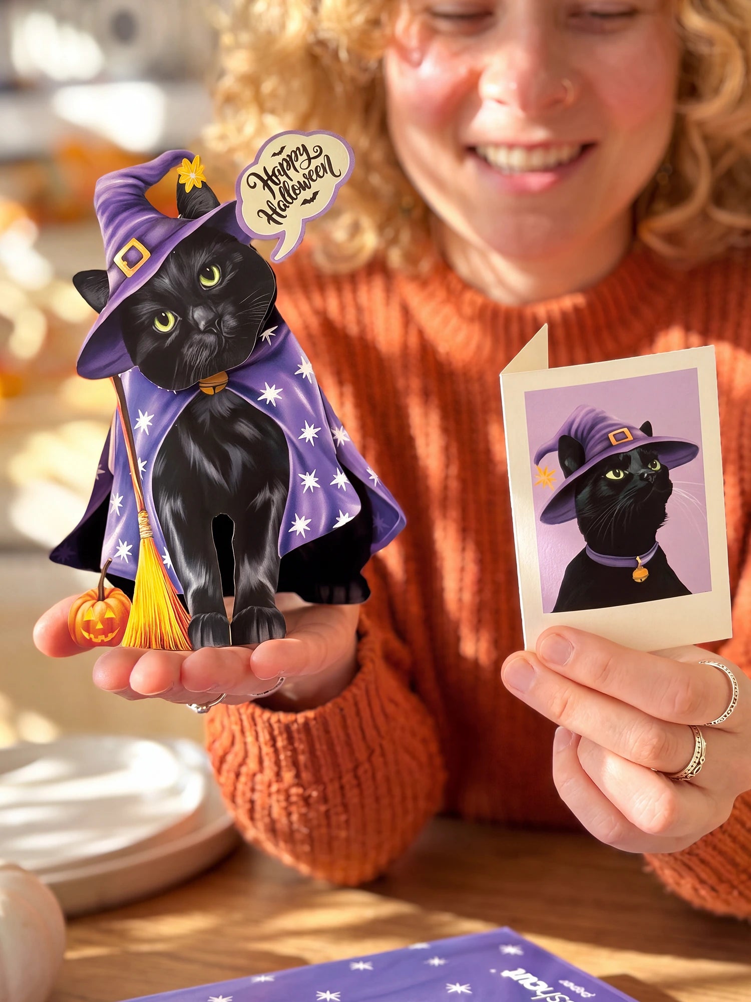 A woman in an orange sweater holds a black cat pop-up card dressed as a witch in a purple hat and cape with a broom, alongside a small 'Happy Halloween' card and a printed photo of the cat.