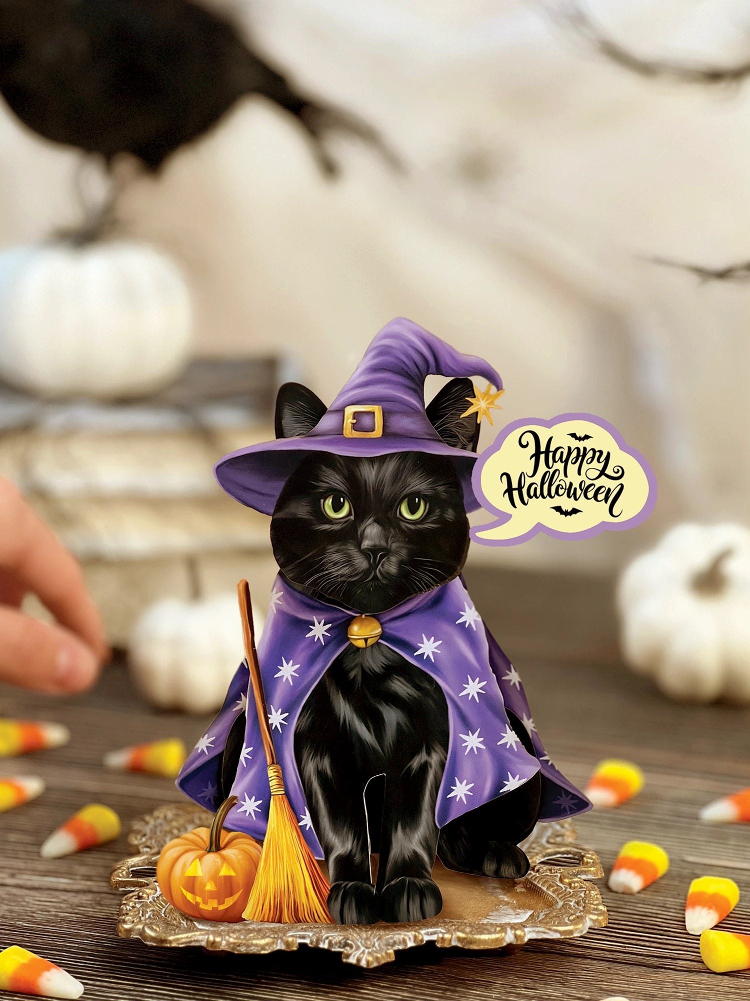 A black cat pop-up card dressed as a witch, wearing a purple pointed hat and matching purple cape with white star patterns, standing on a decorative gold base. A small broomstick rests beside it, and a speech-bubble decoration reads 'Happy Halloween.
