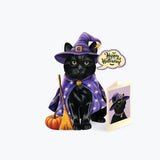 A black cat dressed as a witch wearing a purple hat and cape with white stars, standing beside a small orange pumpkin and broom. A speech bubble says 'Happy Halloween' and an open card shows the same illustration.