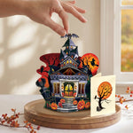A hand is placing a small Halloween-themed decorative house on a round wooden base. The set includes a detailed haunted house with glowing windows, pumpkins, bats, a witch on a broomstick, and a separate card with a spooky tree and full moon, styled on a table near a window.