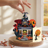 A hand is placing a small Halloween-themed decorative house on a round wooden base. The set includes a detailed haunted house with glowing windows, pumpkins, bats, a witch on a broomstick, and a separate card with a spooky tree and full moon, styled on a table near a window.