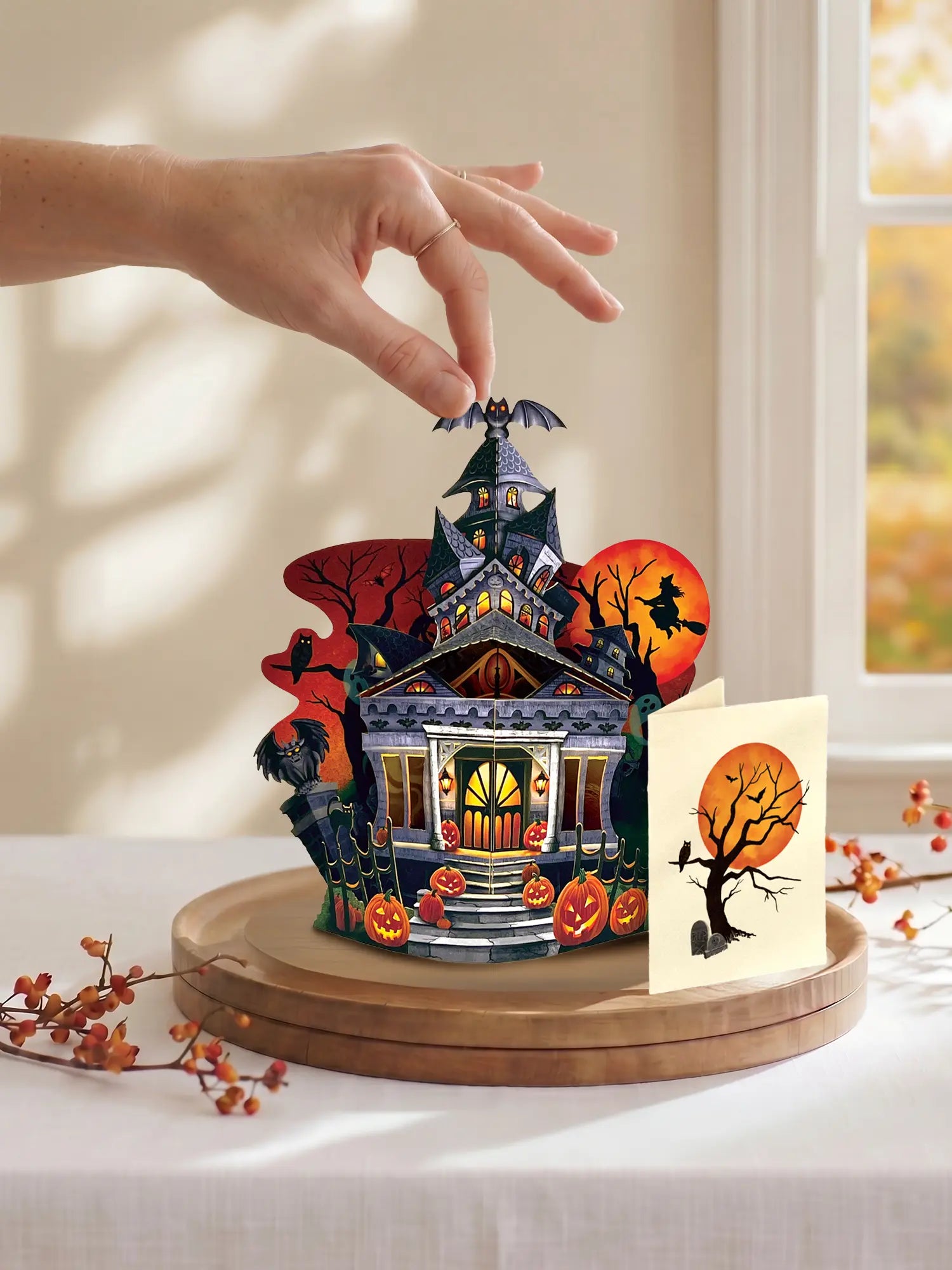 A hand is placing a small Halloween-themed decorative house on a round wooden base. The set includes a detailed haunted house with glowing windows, pumpkins, bats, a witch on a broomstick, and a separate card with a spooky tree and full moon, styled on a table near a window.