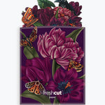 A branded envelope featuring a dark purple background with large magenta flowers, green leaves, orange butterflies, and a black bird perched at the top. The words 'freshcut paper' are printed near the bottom.
