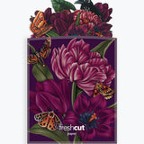 A branded envelope featuring a dark purple background with large magenta flowers, green leaves, orange butterflies, and a black bird perched at the top. The words 'freshcut paper' are printed near the bottom.