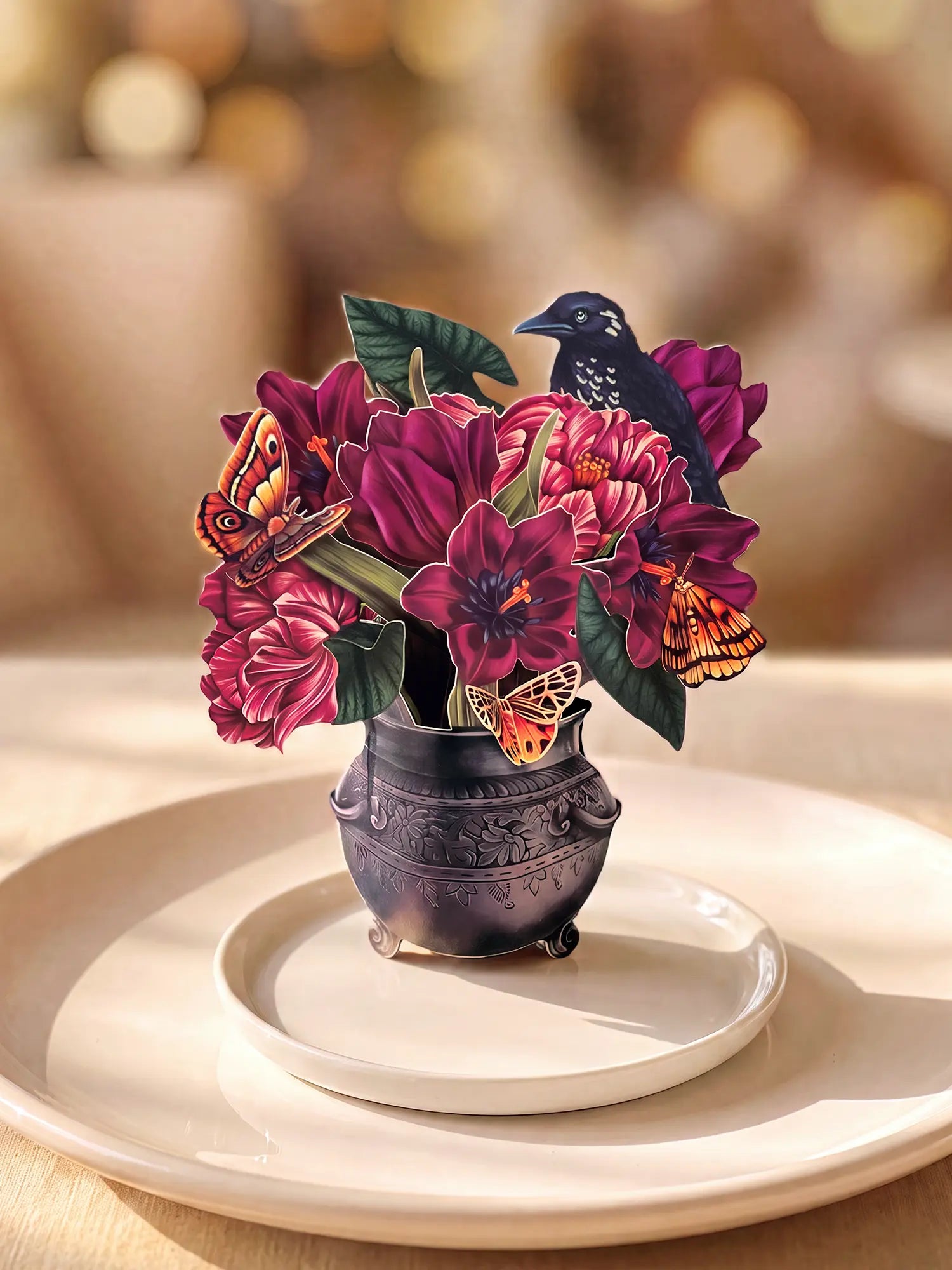 A decorative floral centerpiece in a small carved dark vase, featuring deep magenta flowers, green leaves, orange butterflies, and a black bird, arranged on a white plate against a softly blurred warm background.