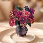 A decorative floral centerpiece in a small carved dark vase, featuring deep magenta flowers, green leaves, orange butterflies, and a black bird, arranged on a white plate against a softly blurred warm background.