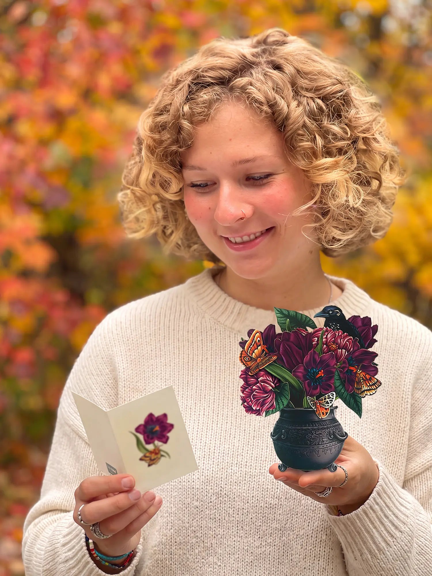 A smiling woman with curly blonde hair looks down at a small card in one hand and holds a small black cauldron containing dark purple flowers and butterflies in the other. She is wearing a cream knit sweater, with a blurred background of autumn foliage in orange and yellow.