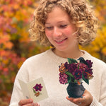A smiling woman with curly blonde hair looks down at a small card in one hand and holds a small black cauldron containing dark purple flowers and butterflies in the other. She is wearing a cream knit sweater, with a blurred background of autumn foliage in orange and yellow.