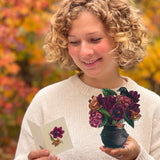 A smiling woman with curly blonde hair looks down at a small card in one hand and holds a small black cauldron containing dark purple flowers and butterflies in the other. She is wearing a cream knit sweater, with a blurred background of autumn foliage in orange and yellow.