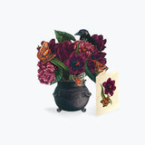 A decorative illustration of a black cauldron filled with dark purple flowers, green leaves, orange butterflies, and a black bird, shown beside a small cream card with a matching floral design on a light gray background.