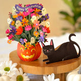 Halloween paper floral arrangement in a carved orange pumpkin pot with colorful flowers, ghost accents, and a purple 'Happy Halloween!' banner, displayed on a wooden stand beside a black cat card silhouette with white pumpkins in the foreground.