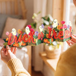 A person holds a colorful 3D pop-up paper banner spelling 'trick or treat' with cartoon ghosts, pumpkins, and flowers in a warm, softly lit room.