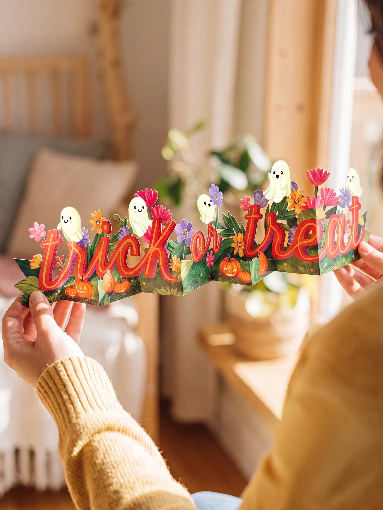 A person holds a colorful 3D pop-up paper banner spelling 'trick or treat' with cartoon ghosts, pumpkins, and flowers in a warm, softly lit room.