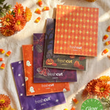 A styled flat-lay of branded envelope packaging of each mini with Halloween designs, arranged on a cream fabric background with orange flowers and candy corn.