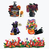 A set of five Halloween-themed decorative items arranged on a plain white background: a haunted house lantern, a black cauldron with dark flowers, a black cat figurine with a witch hat and broom, a jack-o'-lantern planter filled with multicolored flowers and a black cat, and a long garland reading “trick or treat” with ghosts and flowers.
