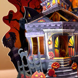 A 3D papercraft Halloween house decoration with carved jack-o'-lantern pumpkins, a black cat on a fence, a spooky tree silhouette, and a haunted house with glowing orange windows.