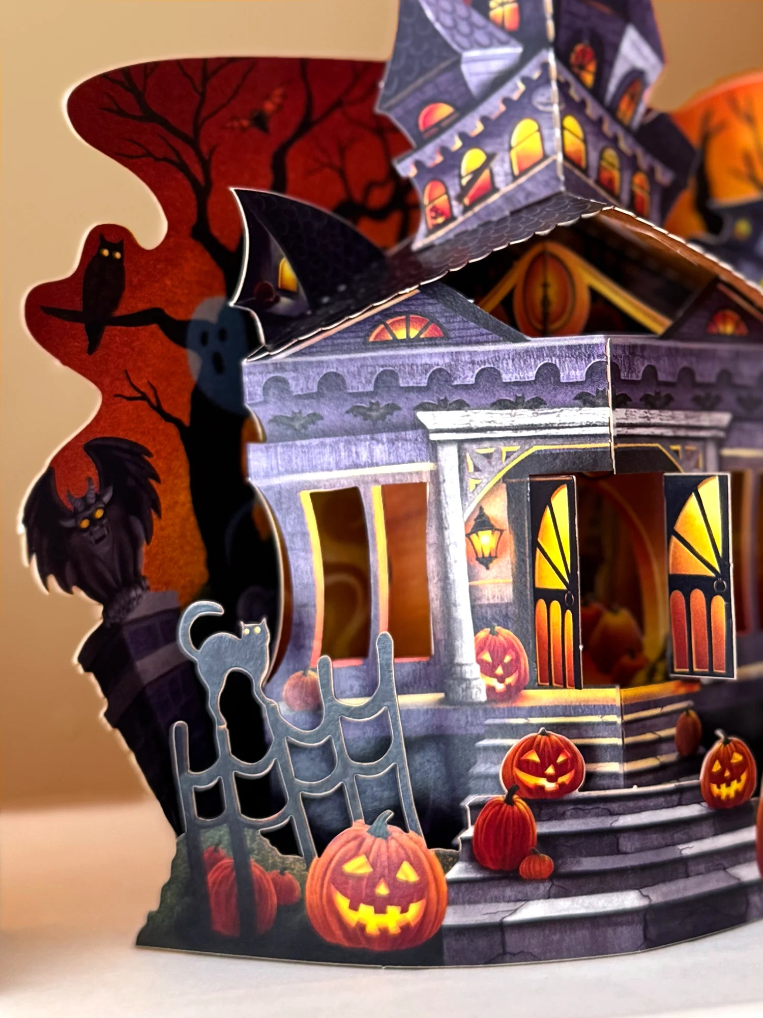 A 3D papercraft Halloween house decoration with carved jack-o'-lantern pumpkins, a black cat on a fence, a spooky tree silhouette, and a haunted house with glowing orange windows.