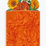 An orange floral envelope with a pop up bouquet coming out of the top with layered orange flowers, yellow sunflowers, and green leaves, shown on a light gray background. The brand name 'freshcut paper' is printed near the bottom.