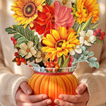 A person in a cream knit sweater holds a vibrant paper floral arrangement in a ribbed orange pumpkin-shaped vase. The bouquet features bright yellow sunflowers, red and pink flowers, white blossoms, and green leaves with small red berries.