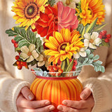A person in a cream knit sweater holds a vibrant paper floral arrangement in a ribbed orange pumpkin-shaped vase. The bouquet features bright yellow sunflowers, red and pink flowers, white blossoms, and green leaves with small red berries.