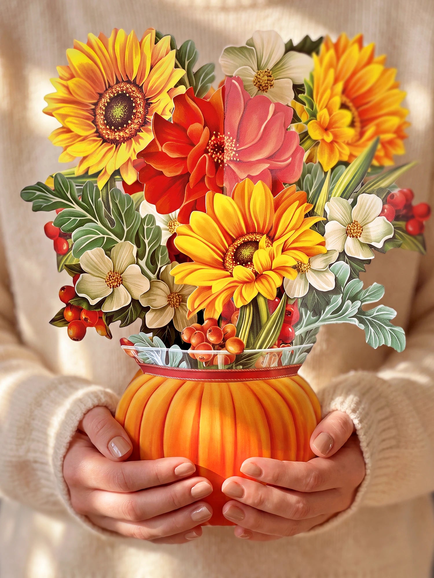A person in a cream knit sweater holds a vibrant paper floral arrangement in a ribbed orange pumpkin-shaped vase. The bouquet features bright yellow sunflowers, red and pink flowers, white blossoms, and green leaves with small red berries.
