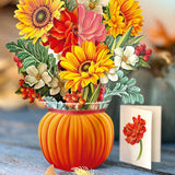 A decorative paper pop-up floral arrangement in a ribbed orange pumpkin-shaped vase, featuring large yellow sunflowers, red and pink blossoms, white flowers, green leaves, and small red berries, set on a rustic wooden surface with a small card beside it.
