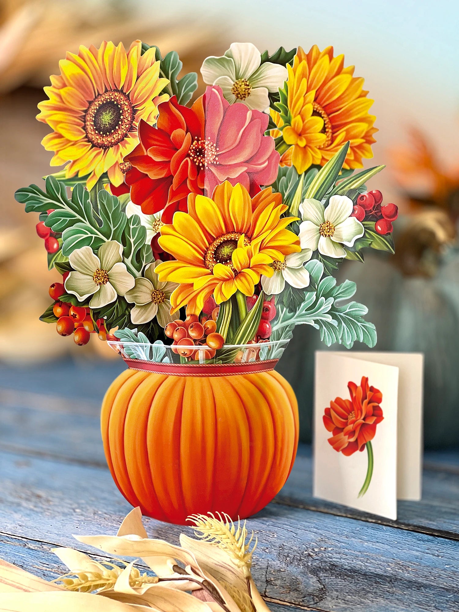 A decorative paper pop-up floral arrangement in a ribbed orange pumpkin-shaped vase, featuring large yellow sunflowers, red and pink blossoms, white flowers, green leaves, and small red berries, set on a rustic wooden surface with a small card beside it.