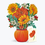 A floral paper pop-up arrangement in a ribbed orange pumpkin-shaped vase, featuring yellow and orange flowers with green leaves and small red berries, alongside a small card with a matching flower illustration, on a light gray background.