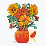 A floral paper pop-up arrangement in a ribbed orange pumpkin-shaped vase, featuring yellow and orange flowers with green leaves and small red berries, alongside a small card with a matching flower illustration, on a light gray background.