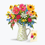 A vibrant mixed flower bouquet in a white ceramic pitcher with a green leaf pattern, accompanied by a small matching flower card on a clean white background.