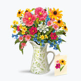A vibrant mixed flower bouquet in a white ceramic pitcher with a green leaf pattern, accompanied by a small matching flower card on a clean white background.