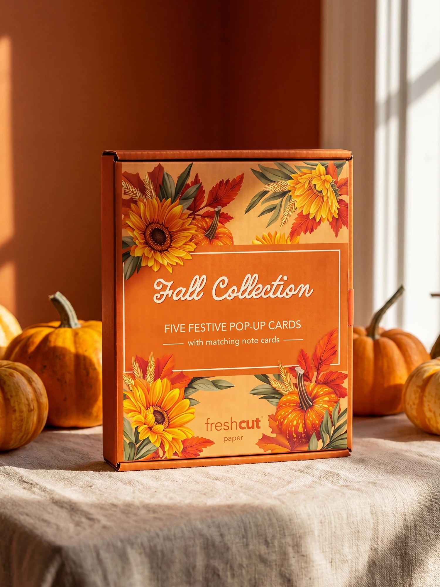 A boxed card set labeled 'Fall Collection' stands on a tablecloth in a styled autumn scene with pumpkins and warm sunlight. The box has orange, yellow, and red floral artwork and visible 'freshcut paper' logo branding.