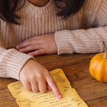 A child in a beige knit sweater points at a yellow autumn-themed scavenger hunt card on a wooden table, with a small orange pumpkin nearby.