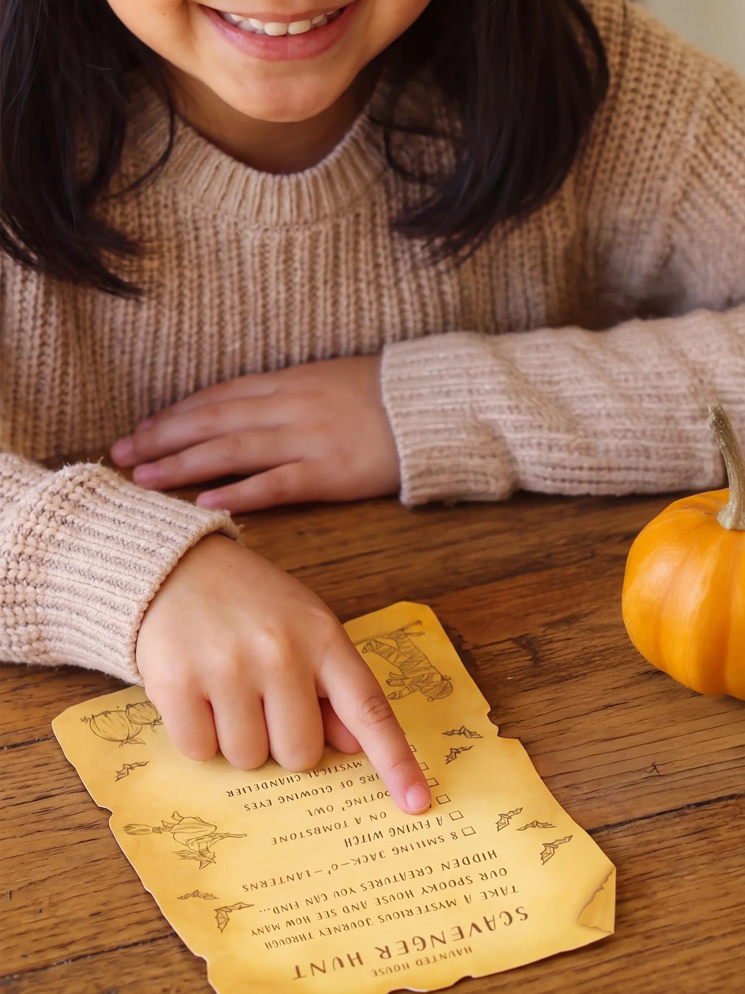 A child in a beige knit sweater points at a yellow autumn-themed scavenger hunt card on a wooden table, with a small orange pumpkin nearby.