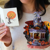 Person holding a pop-up card with a haunted house design and a matching greeting card featuring a spooky tree in front of a glowing orange moon.